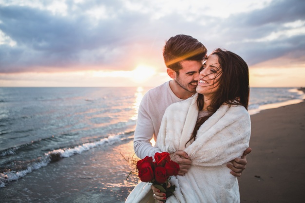 smiling-couple-walking-on-the-beach-with-a-bouquet-of-roses-at-sunset_23-2147595923